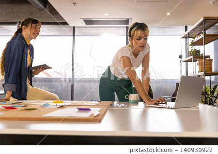 Female colleagues collaborating around large meeting table in office using laptop and tablet Female colleagues collaborating around large meeting table in office using laptop and tablet 140930922