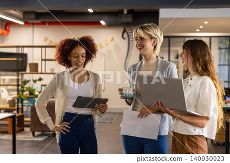 Diverse female coworkers discussing ideas around tablet laptop and mug in modern office lounge Diverse female coworkers discussing ideas around tablet laptop and mug in modern office lounge 140930923
