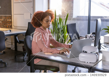 African american woman taking notebook notes while typing on laptop beside vr headset at desk African american woman taking notebook notes while typing on laptop beside vr headset at desk 140930929