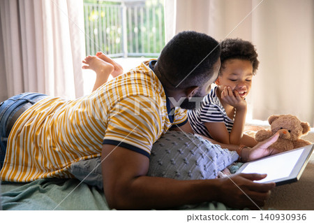 African American father and daughter lying in softly lit bedroom sharing tablet beside teddy bear African American father and daughter lying in softly lit bedroom sharing tablet beside teddy bear 140930936