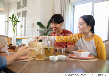 Asian family sitting in dining room sharing meal from steamer basket with iced tea pitcher Asian family sitting in dining room sharing meal from steamer basket with iced tea pitcher 140930937