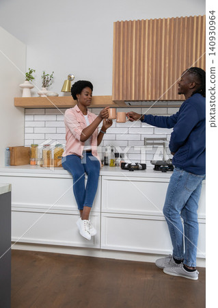 African american couple clinking matching mugs on kitchen countertop next to cooktop and glass jars African american couple clinking matching mugs on kitchen countertop next to cooktop and glass jars 140930964
