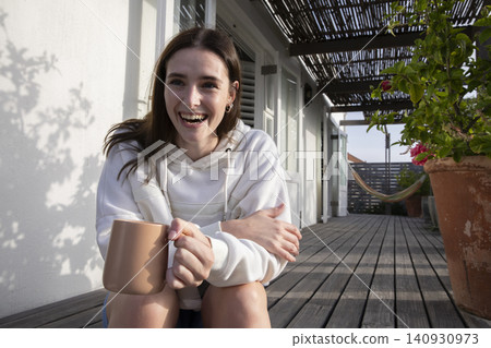 Woman holding large ceramic mug and smiling on wooden veranda under slatted pergola, copy space 140930973