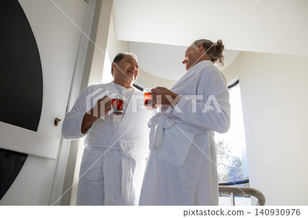 Senior couple standing by balcony railing in spa suite holding mugs of tea wearing bathrobes Senior couple standing by balcony railing in spa suite holding mugs of tea wearing bathrobes 140930976