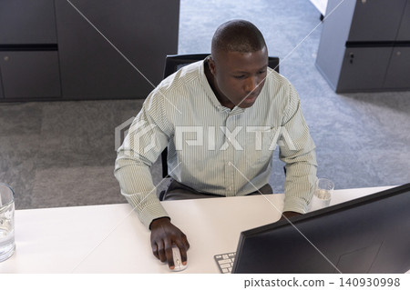 African american man in business attire sitting at office desk using computer with water glasse African american man in business attire sitting at office desk using computer with water glasse 140930998