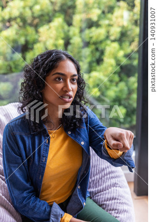 South asian woman in denim jacket sitting on chair near window pointing toward garden South asian woman in denim jacket sitting on chair near window pointing toward garden 140931007