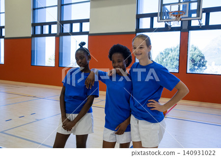Diverse teenage female teammates standing on basketball court inside gym with hoop overhead Diverse teenage female teammates standing on basketball court inside gym with hoop overhead 140931022