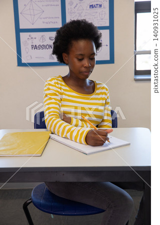 African american female teen writing in notebook with pencil at desk in classroom African american female teen writing in notebook with pencil at desk in classroom 140931025