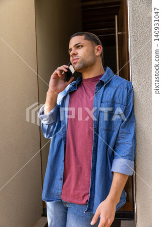 Man in twenties leaning against corridor wall wearing denim shirt talking on smartphone Man in twenties leaning against corridor wall wearing denim shirt talking on smartphone 140931047