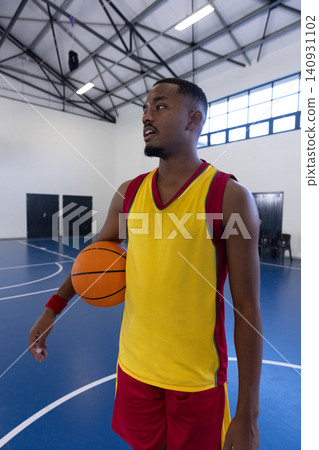 African american adult male standing on blue court in court under metal ceiling, holding basketball African american adult male standing on blue court in court under metal ceiling, holding basketball 140931102