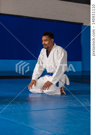African american male martial artist kneeling on mats in dojo in white gi, blue belt African american male martial artist kneeling on mats in dojo in white gi, blue belt 140931110