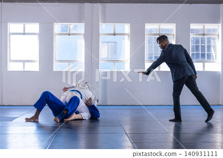 Diverse martial artists grappling in gis on dojo mats with referee signaling by windows Diverse martial artists grappling in gis on dojo mats with referee signaling by windows 140931111