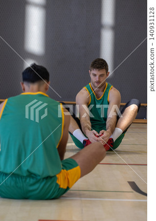 Diverse male teammates stretching on gym floor gripping red sneakers in uniforms Diverse male teammates stretching on gym floor gripping red sneakers in uniforms 140931128