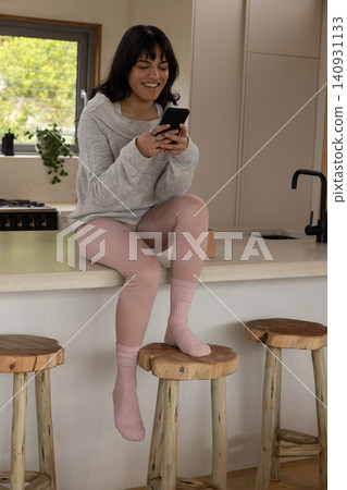 Woman sitting on kitchen island scrolling smartphone beside stove, potted plant on window sill Woman sitting on kitchen island scrolling smartphone beside stove, potted plant on window sill 140931133