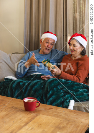 Senior couple wearing Santa hats sharing wrapped gift on sofa in living room holding frothy mug Senior couple wearing Santa hats sharing wrapped gift on sofa in living room holding frothy mug 140931160