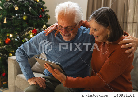 Senior couple smiling while holding tablet on sofa in living room with Christmas tree decorations Senior couple smiling while holding tablet on sofa in living room with Christmas tree decorations 140931161