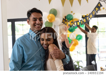 Diverse couple hugging in home living room among green white yellow balloons, pennant garlands Diverse couple hugging in home living room among green white yellow balloons, pennant garlands 140931194