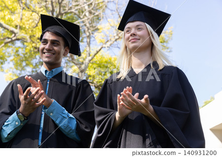Indian man and woman graduates clapping on lawn wearing black gowns, caps, watch Indian man and woman graduates clapping on lawn wearing black gowns, caps, watch 140931200