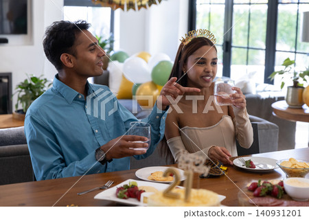 Indian couple toasting birthday with glasses at home table beside cake, girl in tiara, copy space Indian couple toasting birthday with glasses at home table beside cake, girl in tiara, copy space 140931201