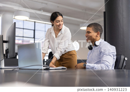 Diverse coworkers pointing at laptop screen in open-plan office with headphones, monitor Diverse coworkers pointing at laptop screen in open-plan office with headphones, monitor 140931223