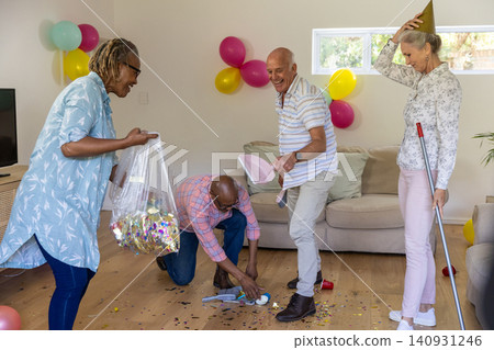 Diverse senior friends tidying living room after party, collecting confetti with dustpan Diverse senior friends tidying living room after party, collecting confetti with dustpan 140931246