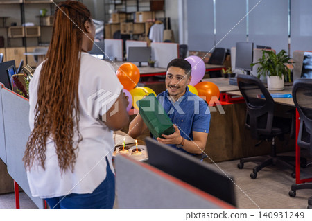 Diverse coworkers handing green gift bag in office beside monitors with balloons, cupcakes Diverse coworkers handing green gift bag in office beside monitors with balloons, cupcakes 140931249