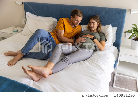 Couple lying on bed touching pregnant belly and sharing glances near nightstands with plant Couple lying on bed touching pregnant belly and sharing glances near nightstands with plant 140931250
