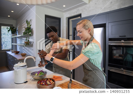 Diverse couple wearing aprons seasoning salad using salt, pepper grinders at kitchen island Diverse couple wearing aprons seasoning salad using salt, pepper grinders at kitchen island 140931255