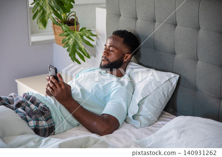 African american man relaxing on bed by headboard in pajamas, scrolling smartphone African american man relaxing on bed by headboard in pajamas, scrolling smartphone 140931262