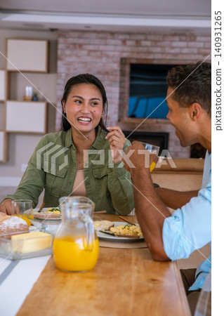 Diverse couple sitting at home sharing plates of scrambled eggs on table with juice jug Diverse couple sitting at home sharing plates of scrambled eggs on table with juice jug 140931265