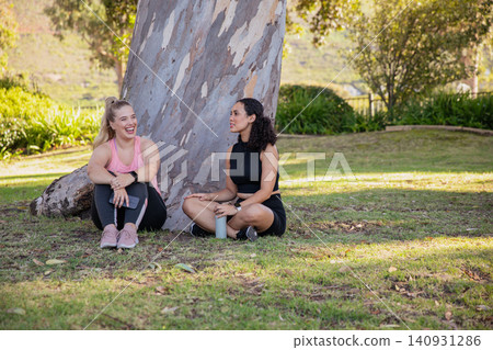 Diverse female friends in activewear sitting by tree in park holding smartphone, water bottle Diverse female friends in activewear sitting by tree in park holding smartphone, water bottle 140931286