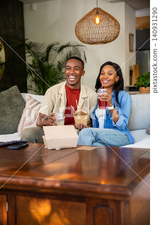 African American couple sitting on pale sofa in living room, holding take-out and sipping drinks African American couple sitting on pale sofa in living room, holding take-out and sipping drinks 140931290