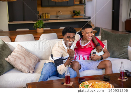 Diverse male friends celebrating and watching match on sofa in living room with pizza, soccer ball Diverse male friends celebrating and watching match on sofa in living room with pizza, soccer ball 140931296