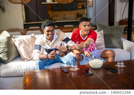 Diverse male friends holding game controllers and grabbing popcorn from coffee table in living room 140931298
