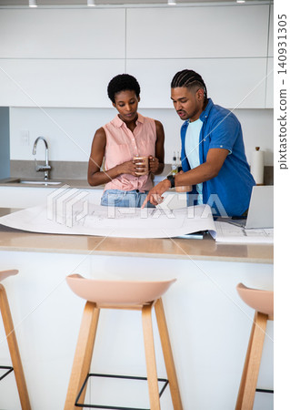 Diverse couple standing at kitchen island pointing at blueprint while holding ceramic mug Diverse couple standing at kitchen island pointing at blueprint while holding ceramic mug 140931305