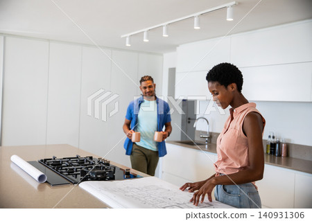 Diverse couple reviewing blueprints and carrying coffee mugs on kitchen island, copy space Diverse couple reviewing blueprints and carrying coffee mugs on kitchen island, copy space 140931306