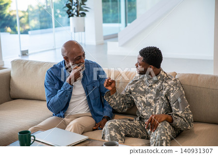 African american father bumping fists with soldier son at home by coffee table with laptop African american father bumping fists with soldier son at home by coffee table with laptop 140931307