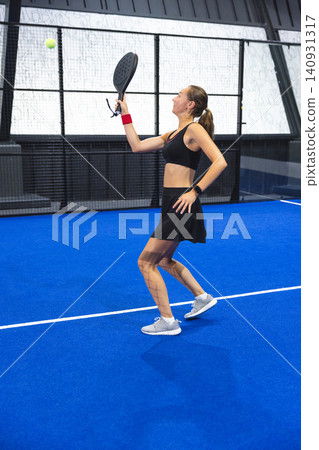 Female athlete hitting yellow ball with padel racket on blue padel court wearing black sportswear Female athlete hitting yellow ball with padel racket on blue padel court wearing black sportswear 140931317