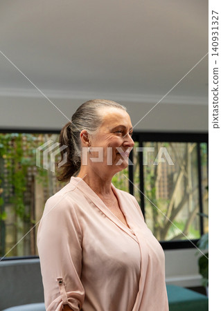 Mature woman standing by black windows in living room, wearing pale pink blouse, teal sofa visible Mature woman standing by black windows in living room, wearing pale pink blouse, teal sofa visible 140931327