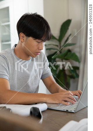 Asian adult male wearing gray T-shirt, working on silver laptop at table at home with headset Asian adult male wearing gray T-shirt, working on silver laptop at table at home with headset 140931328