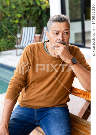 Senior man sitting on wooden bench at poolside, wearing rust sweater, with watch and ring visible 140931330