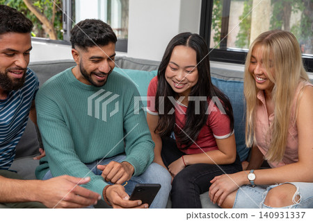 Diverse friends sitting on gray couch in living room, sharing smartphone and pointing at screen Diverse friends sitting on gray couch in living room, sharing smartphone and pointing at screen 140931337