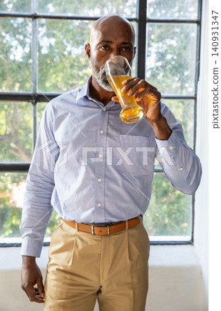 Senior African American man sipping from tall glass of orange-juice at multi-pane window in shirt Senior African American man sipping from tall glass of orange-juice at multi-pane window in shirt 140931347