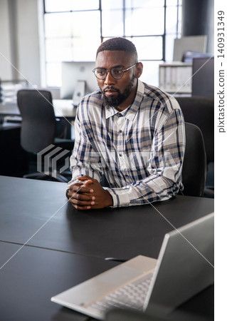 Adult African American man sitting facing laptop at office table, wearing plaid shirt and glasses 140931349