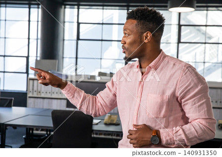 Adult African American male in light pink shirt standing, pointing in office with desks, wristwatch Adult African American male in light pink shirt standing, pointing in office with desks, wristwatch 140931350