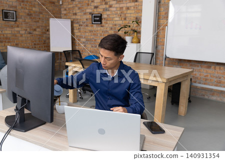Adult Asian man pouring coffee from carafe into mug wearing navy shirt at desk with monitor Adult Asian man pouring coffee from carafe into mug wearing navy shirt at desk with monitor 140931354