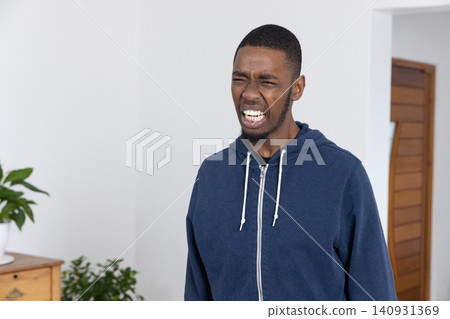 Adult African American man grimacing with eyes closed, standing in room in navy hoodie near dresser Adult African American man grimacing with eyes closed, standing in room in navy hoodie near dresser 140931369