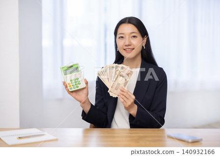 一位年輕的日本女商人對著鏡頭微笑,手裡拿著計算機和一張10,000日圓的鈔票。 一位年輕的日本女商人對著鏡頭微笑,手裡拿著計算機和一張10,000日圓的鈔票。 140932166