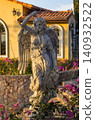 Classical winged angel statue holding grapes among bougainvillea with a Mediterranean-style building in warm evening light 140932522