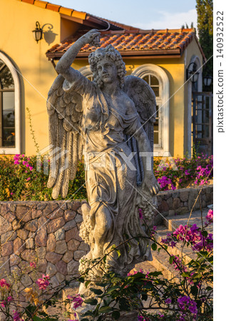 Classical winged angel statue holding grapes among bougainvillea with a Mediterranean-style building in warm evening light Classical winged angel statue holding grapes among bougainvillea with a Mediterranean-style building in warm evening light 140932522